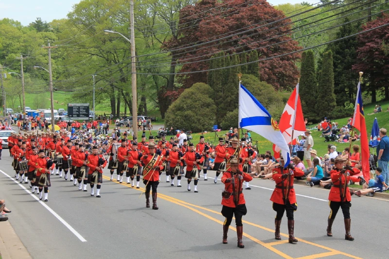 RCMP officers in red serge uniforms march in a parade carrying the Nova Scotia, Canadian, and Red Ensign flags, followed by an RCMP pipes and drums band.